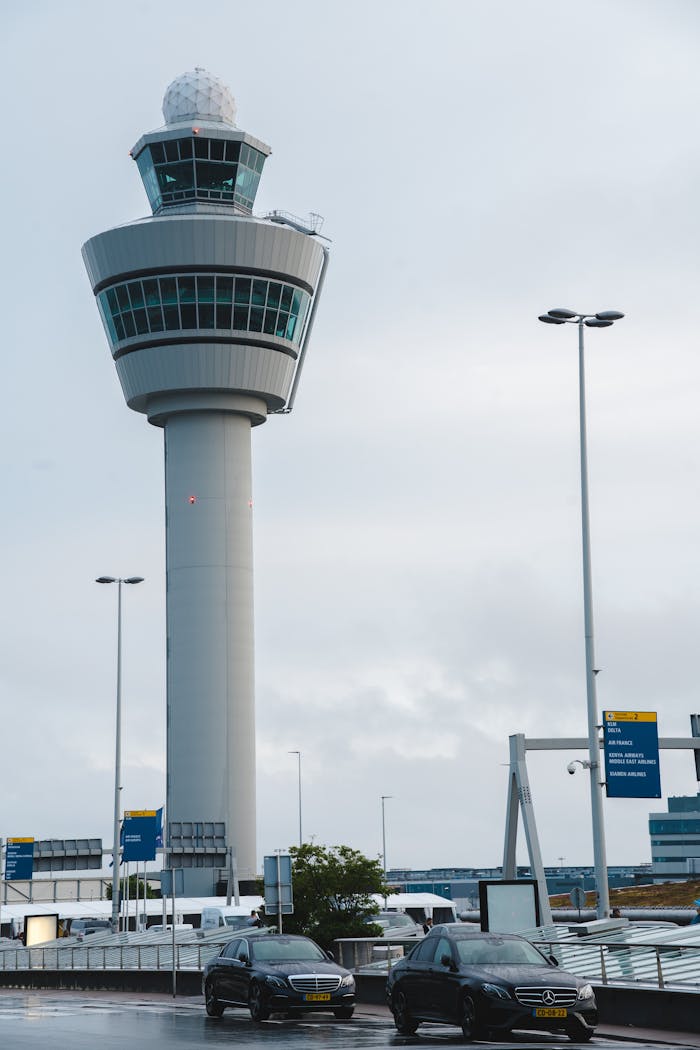 Iconic Schiphol Airport control tower under cloudy skies, with vehicles in the foreground.
